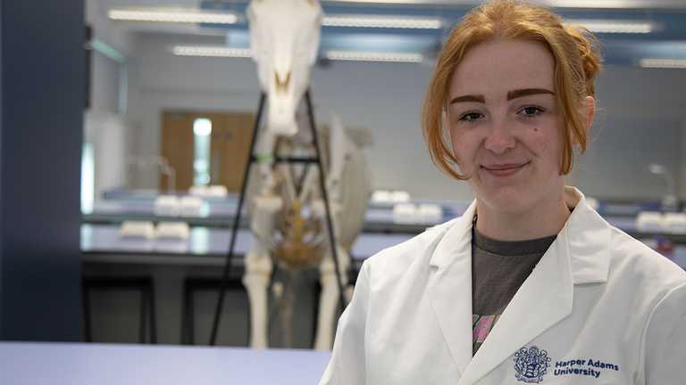 A woman - Martha Stokes - in a white lab coat with a hore skeleton behind her.