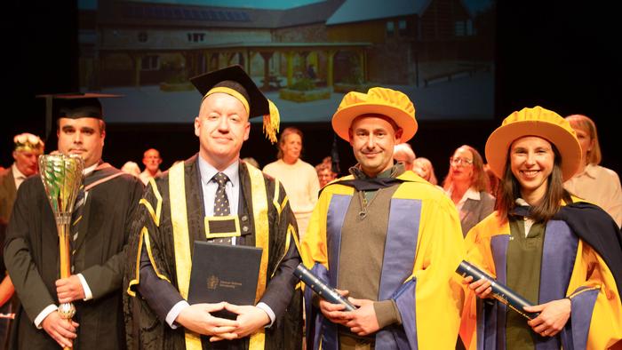 Mace Bearer Dr Jonathan Cooper, Professor Ken Sloan, and new Honorary Graduates Ben and Charlotte Hollins onstage at Theatre Severn