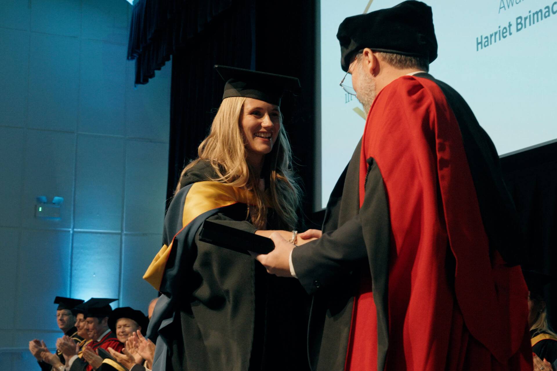 Harriet wearing graduation gown and cap accepting an award from Professor Michael Lee.
