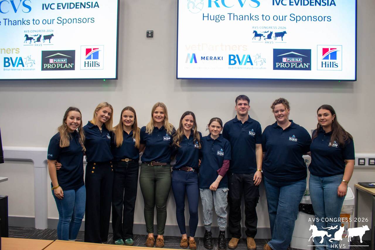 Photograph of nine people standing together in a line, two large screens behind them with various logos displayed.